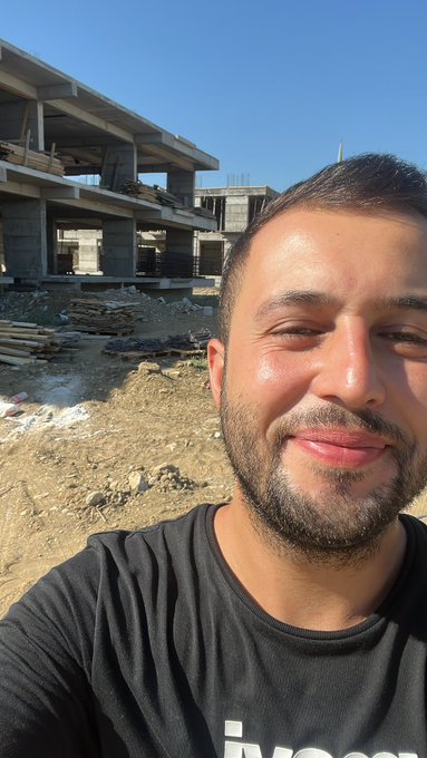 A bearded man takes a selfie in front of an unfinished concrete building under construction, set against a clear blue sky in a dry, rural landscape.