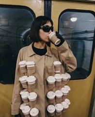 A person with dark hair and sunglasses wearing a trench coat, holding multiple stacked coffee cups and drinking from one, posing in front of a yellow vehicle.