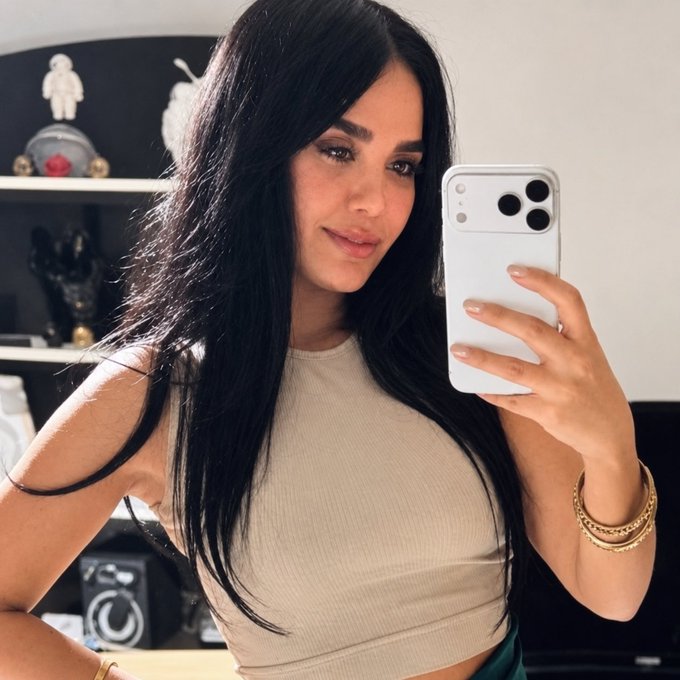 A woman with long black hair takes a mirror selfie with a white smartphone, wearing a beige crop top and gold bracelets, in a room with shelves displaying figurines.