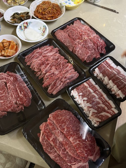 An overhead view of multiple black trays filled with various cuts of raw marbled meat, accompanied by Korean side dishes, prepared for a Korean barbecue meal.