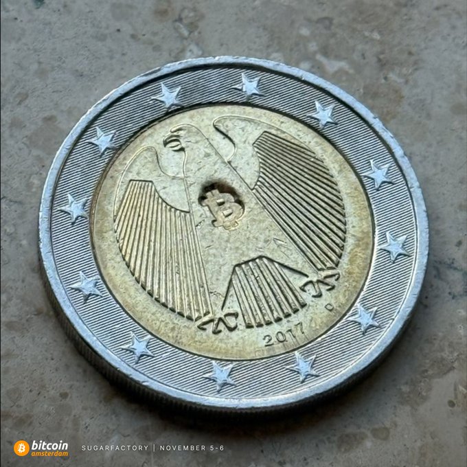 A close-up of a 2017 2 Euro coin featuring the German eagle design, with 'Bitcoin Amsterdam' and 'Sugarfactory | November 5-6' text in the corner.
