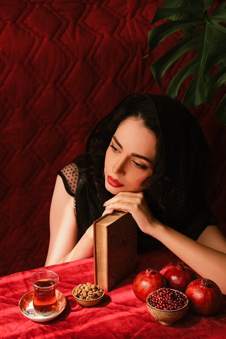 A woman with long dark hair and red lipstick rests her chin on a wooden block on a red velvet table, with a glass of tea, pomegranates, and a bowl of nuts arranged in front of her against a dark red backdrop.