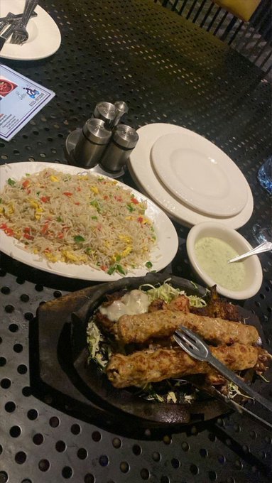 A restaurant meal on a dark textured table featuring a plate of vegetable fried rice, a sizzling platter of grilled meat skewers over cabbage, and a small dish of green sauce.
