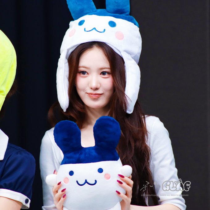 A young woman with long brown hair wearing a white Cinnamoroll character hat and holding a matching Cinnamoroll plush toy, smiling at a fan event.