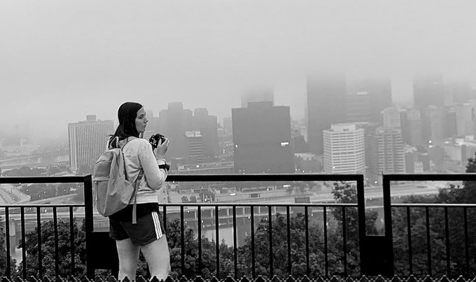 A black and white photograph of a woman wearing a backpack and holding a camera, looking out over a foggy city skyline from a viewing platform.