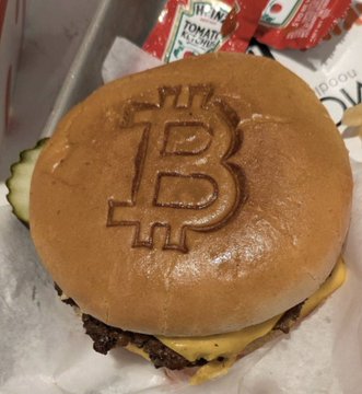 A Steak 'n Shake burger features the iconic Bitcoin "₿" symbol branded onto its bun, accompanied by Heinz ketchup and what appears to be tomato ketchup in the background, visually celebrating the chain's recent embrace of Bitcoin Principles and reported success in surpassing McDonald's in store sales.