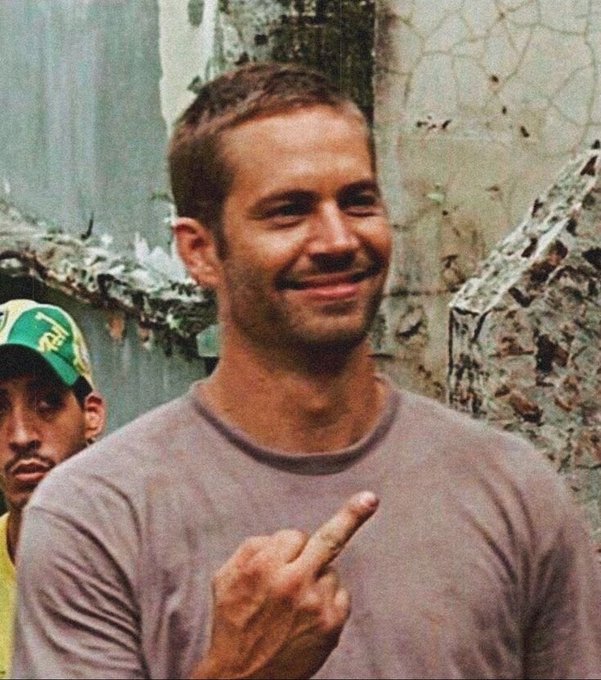 A man with short brown hair, wearing a grey t-shirt, smiles and gestures with his middle finger, standing in front of a dilapidated wall with another man in a green cap partially visible behind him.
