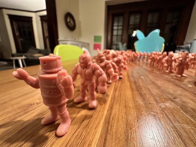A long row of pink muscular action figures is lined up on a wooden floor, with a yellow rubber duck and a blue cat-shaped object visible in the background.