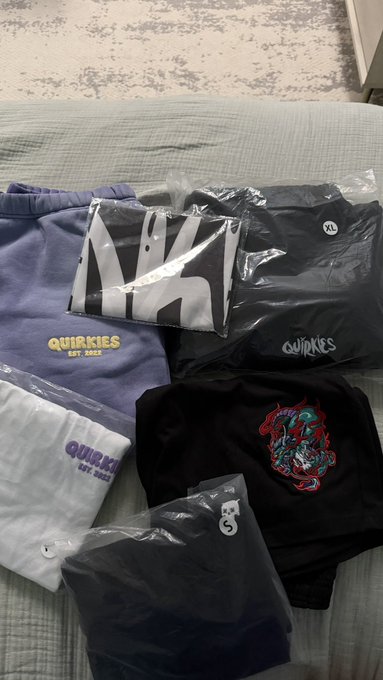 A collection of clothing items including hoodies and a t-shirt with 'QUIRKIES EST 2022' branding laid out on a bed in plastic packaging with XL size tags.