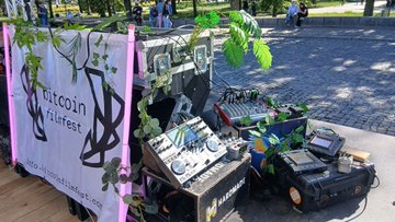 An outdoor display at the 'Bitcoin Frühfest' features electronic equipment including a small robotic vehicle and various devices, with a banner reading 'bitcoin-frühfest' and a Bitcoin logo.