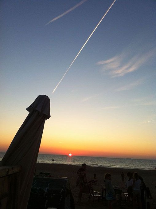 At sunset on a beach, contrails streak across the sky, as people gather near an umbrella. The image captures a serene moment that complements the post's greeting and call to "hunt for alphas," perhaps symbolizing new opportunities or explorations.