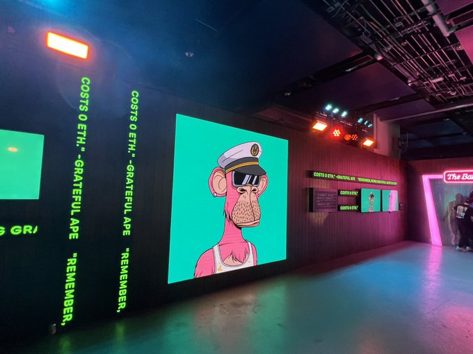 A gallery or exhibition space displays a large neon-lit Bored Ape Yacht Club NFT artwork with text reading 'COSTS 9.5 ETH, "GRATEFUL APE," REMEMBER,' and 'The Bak' visible in the background.