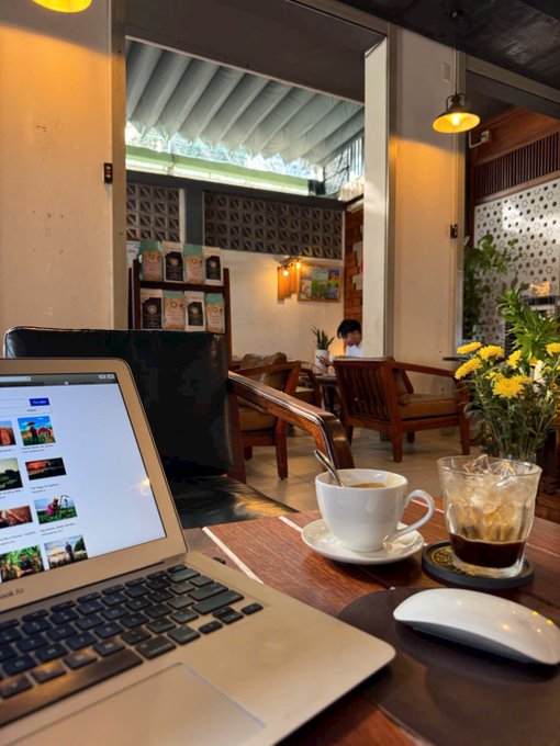 The image shows a laptop computer sitting on top of a wooden table, with a cup of coffee next to it. The background reveals that this scene takes place in an office or workspace setting. A person can be seen seated at another desk in the room, possibly working or attending a meeting. There are also chairs and potted plants present in the space, adding to its overall ambiance.