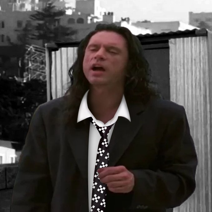 A man with long dark hair wearing a black suit, white shirt, and a black-and-white checkered tie, appearing to sing or speak outdoors.