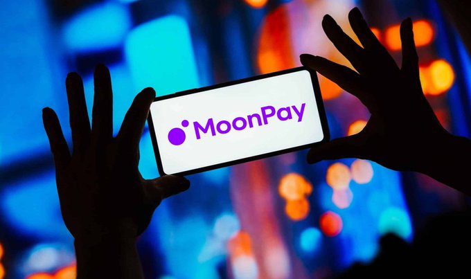 Silhouetted hands holding a smartphone displaying the MoonPay logo against a blurred blue and orange city background.