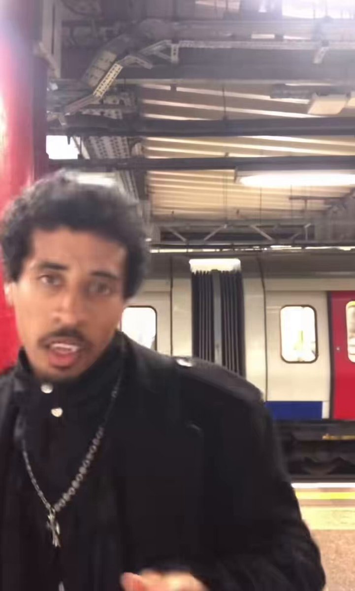 The image shows a man wearing a black jacket, standing next to a train at a station. He has his mouth open as if he's speaking or making a statement. The background of the photo features a train with red doors, indicating that this scene takes place in an urban setting where public transportation is available. The caption provided by the image suggests that the man might be discussing something related to the train service or expressing dissatisfaction about it.