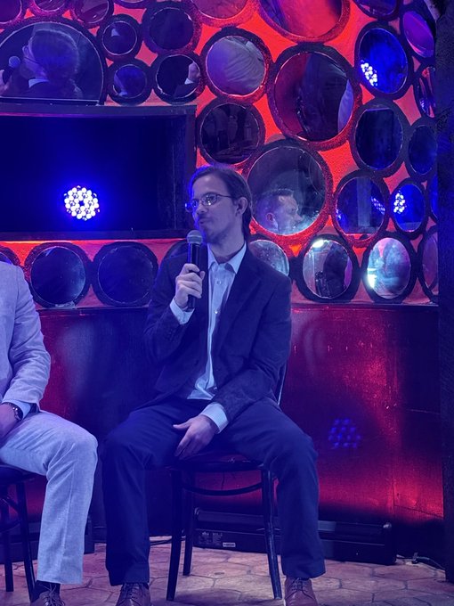 A man in a dark suit and glasses speaks into a microphone while seated on a stage with a distinctive backdrop of circular mirrors illuminated by red and blue lights.