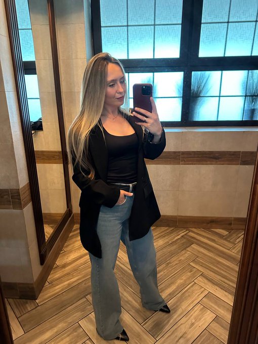 A woman with long blonde hair poses in a mirror selfie wearing a black blazer, black top, and light blue jeans with a belt, standing in a tiled bathroom with a large frosted window. The polished outfit complements the upbeat weekend greeting, presenting a confident, stylish start to the day for her Web3/CT audience.
