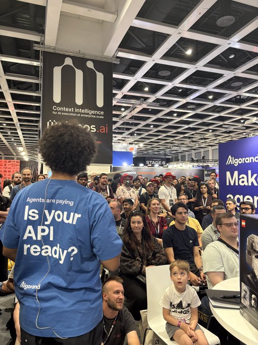 A person wearing a blue t-shirt with 'Agents are paying Is your API ready? Algorand' standing before a crowd at what appears to be a tech conference.