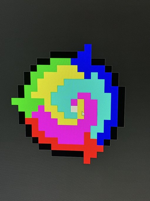 A 3D-rendered pixelated spiral or pinwheel design composed of green, yellow, pink, red, and blue blocks against a dark grey background.