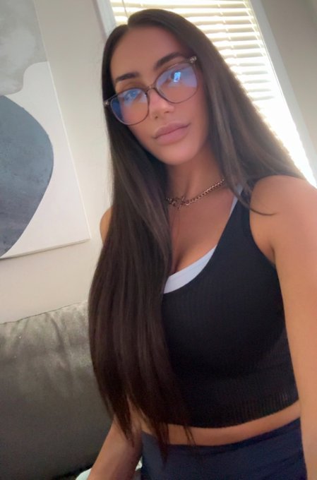 A woman with long dark hair, blue-tinted glasses, and a chain necklace takes a selfie while wearing a black tank top.