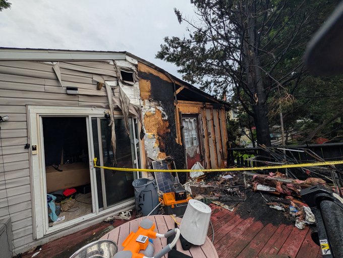 The aftermath of a house fire showing significant structural damage to a residential home, with caution tape strung across a debris-filled wooden deck.