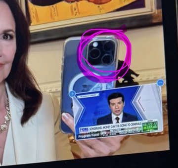 The image shows a woman holding up a cell phone displaying an advertisement or news segment featuring a man wearing a suit and tie. The text in the image reads "XRP - Its COMING REAL SOON («IN A COUPLE OF MINUTES») Can’t say they didn’t tell us; Also: Dr. Judy Shelton is well-known for advocating a return to the GOLD STANDARD to limit government currency manipulation and ensure «honest» money". The woman's reflection in the cell phone screen adds an interesting visual element to the scene, as it shows her face along with the advertisement or news segment.