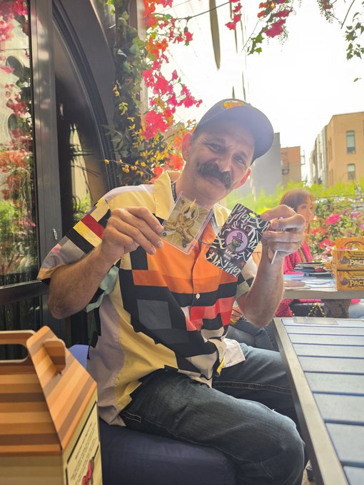 A man wearing a colorful geometric-patterned shirt and a Red Bull cap sits outdoors at a table, holding up two collectible cards and smiling.