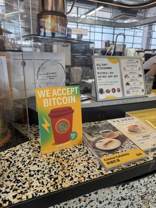 A counter at a Cha Cha coffee shop displaying a colorful 'WE ACCEPT BITCOIN' sign next to a menu showing 'Crema Catalana' for $20, with an 'ALL DAY 全日COMBO' menu in the background.