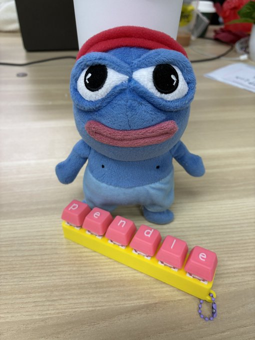 A blue fuzzy plush character with a red beanie and sad expression sitting on a desk behind a yellow keyboard with pink 'pendle' keycaps.