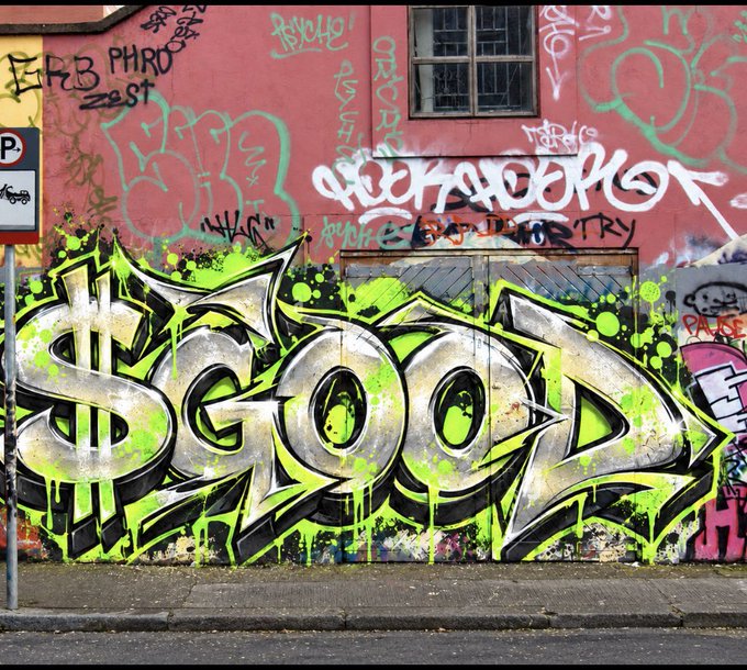 A photograph of urban graffiti on a pinkish-red wall, prominently featuring the word "GOOD" in large stylized letters with black outlines, white fill, and dripping neon green accents, surrounded by other tags and a "P" parking sign.
