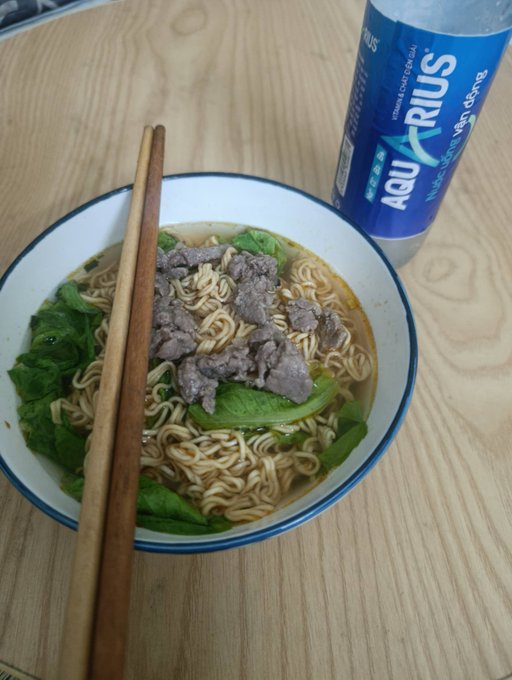 A bowl of instant noodles topped with beef and lettuce sits on a wooden table with chopsticks resting on the bowl, accompanied by a blue Aquarius water bottle.