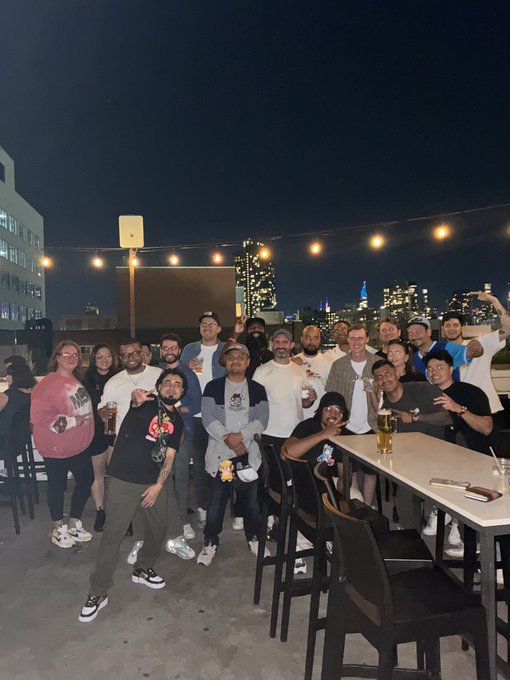 A large group of people posing together for a photo at night on an outdoor rooftop with string lights and a city skyline in the background.