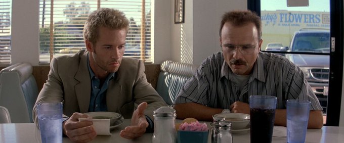 Two men sit at a diner table with coffee cups; a sign reading 'FLOWERS' is visible through the window behind them.