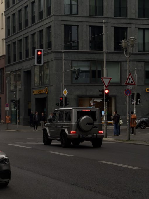 A dark Mercedes-Benz G-Class SUV driving through a city intersection at a red light, passing a Commerzbank branch on the corner.