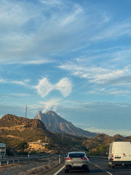 A scenic photograph taken from a car on a highway, showing a rugged mountain peak under a partly cloudy sky, with two vehicles visible ahead.