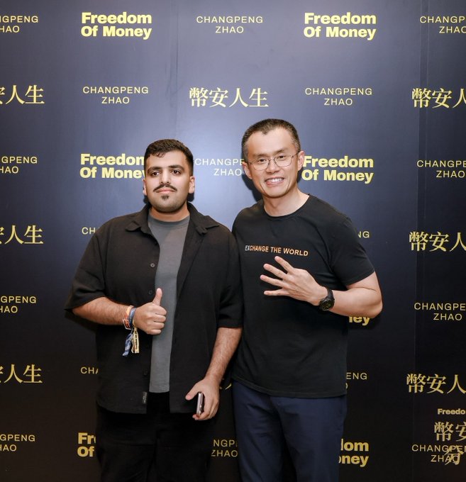 Two men pose in front of a black backdrop branded with "Freedom of Money" and "CHANGPENG ZHAO / 幣安人生" logos, with the man on the right wearing a "Change the World" t-shirt and giving a peace sign. The image captures the Hong Kong event appearance alongside CZ, as mentioned in the accompanying post celebrating the Binance and BNB Chain community.