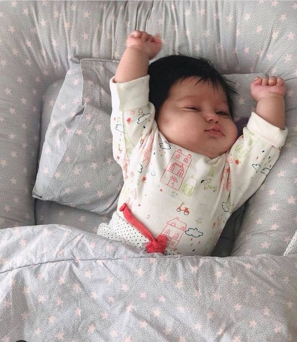 A newborn baby with dark hair stretches both arms above their head while lying on a gray star-patterned bedding and pillow, wearing a white onesie printed with colorful houses, clouds, and bicycles.