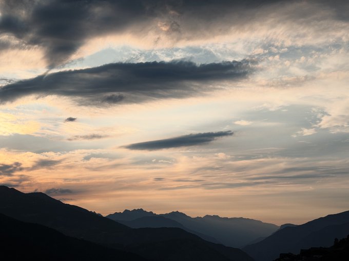 A scenic landscape of silhouetted mountain ranges beneath a dramatic sky filled with layered, wispy clouds at sunset.