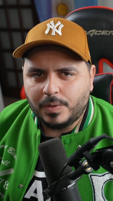 A man wearing a yellow "NY" Yankees cap and a green varsity jacket with the number "10" speaks into a Shure microphone, likely recording a YouTube video. The setup aligns with the post promoting an educational video about the play-to-earn game VikingsValley on his YouTube channel.