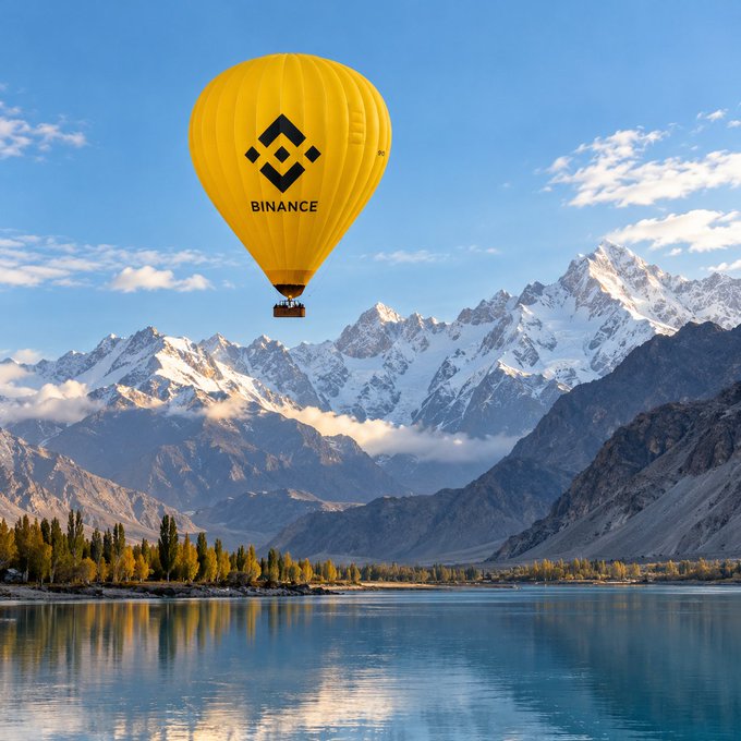 A yellow hot air balloon featuring the Binance logo flies over a scenic landscape of snow-capped mountains and a calm lake reflecting the scenery.