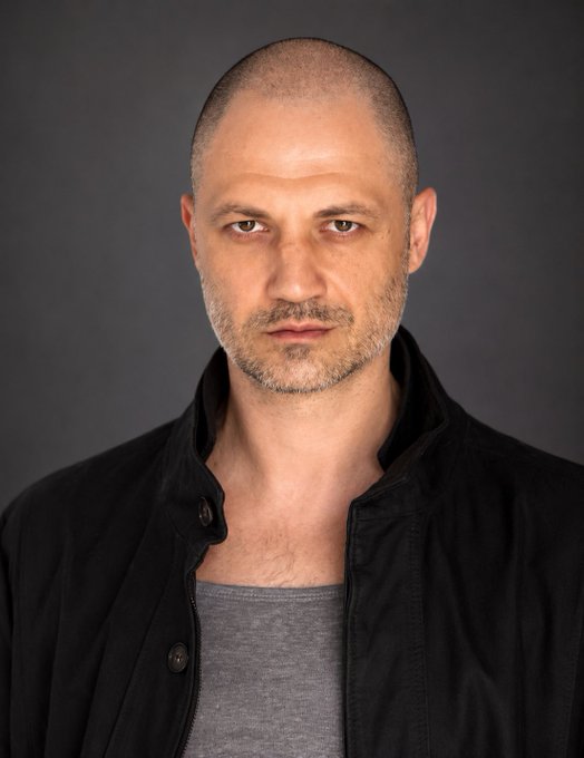 A studio portrait of a bald, middle-aged man with a short beard, wearing a black jacket over a gray shirt against a dark gray background.