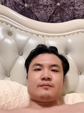 The image shows a man lying down on a bed with his head resting against the headboard of the bed. The man appears to be looking directly at the camera, giving off an impression that he might be posing for a photo or selfie. The background is blurred and out of focus, drawing attention to the main subject - the man in bed.