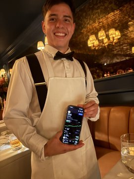 A smiling waiter in a white shirt, black bowtie, and suspenders holds up a smartphone showing a stock chart while standing in an upscale restaurant with chandeliers in the background. The image humorously suggests the waiter is sharing insider stock tips about $CATE/gCATE tokens to skeptical patrons.
