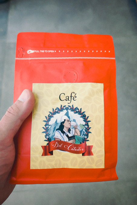 A hand holds an orange coffee bag labeled "Café Del Catador" featuring an illustrated woman with a coffee cup surrounded by a wreath of coffee berries, representing the Colombian coffee mentioned in the post as being received via Cakewallet from EscuelitaBTC.