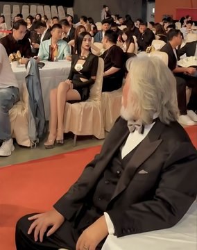 A photograph shows an older man with white hair and a mustache, wearing a black suit, sitting at an event with rows of seated attendees in the background.