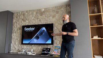 The image shows a man standing in front of a flat screen TV, which displays an advertisement for the Samsung smartphone. The man appears to be giving a presentation or discussing the features of the phone with someone else. There are also several books visible on a nearby surface and a laptop placed next to them.