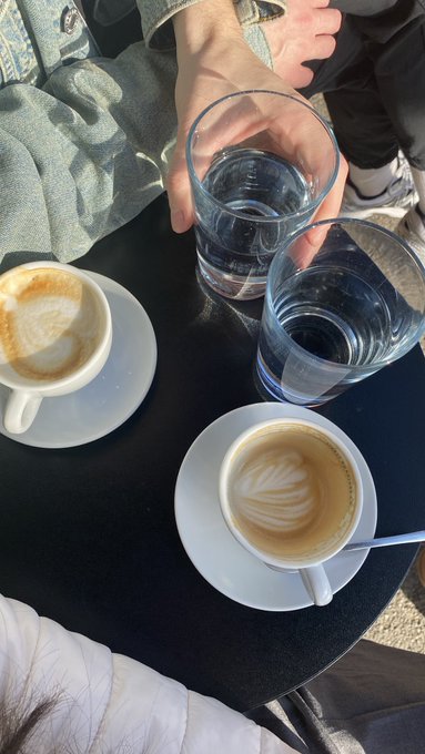 An overhead view of a cafe table with two cups of latte featuring leaf-patterned foam art and two glasses of water, with people's hands and legs visible around the table.