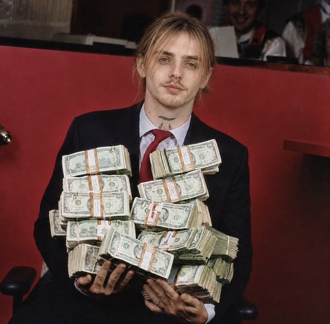 A young man with long blonde hair, a mustache, and a suit holds a large bundle of stacked US dollar bills against a red background.