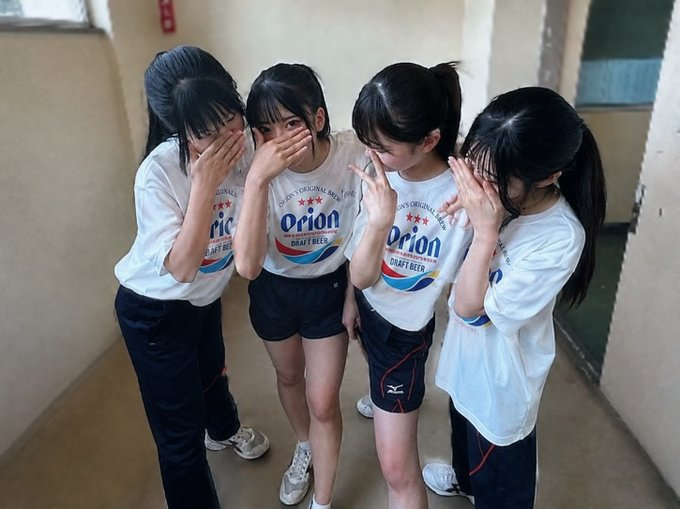 Four young women wearing matching white 'Orion' t-shirts and dark shorts standing together in a hallway, covering their mouths with their hands in a shy pose.